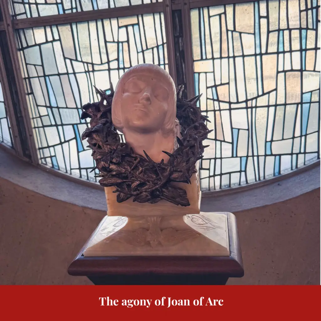 Pale marble bust titled The Agony of Joan of Arc, her head with closed eyes encircled by a bronze wreath of thorns, set against a stained-glass window.