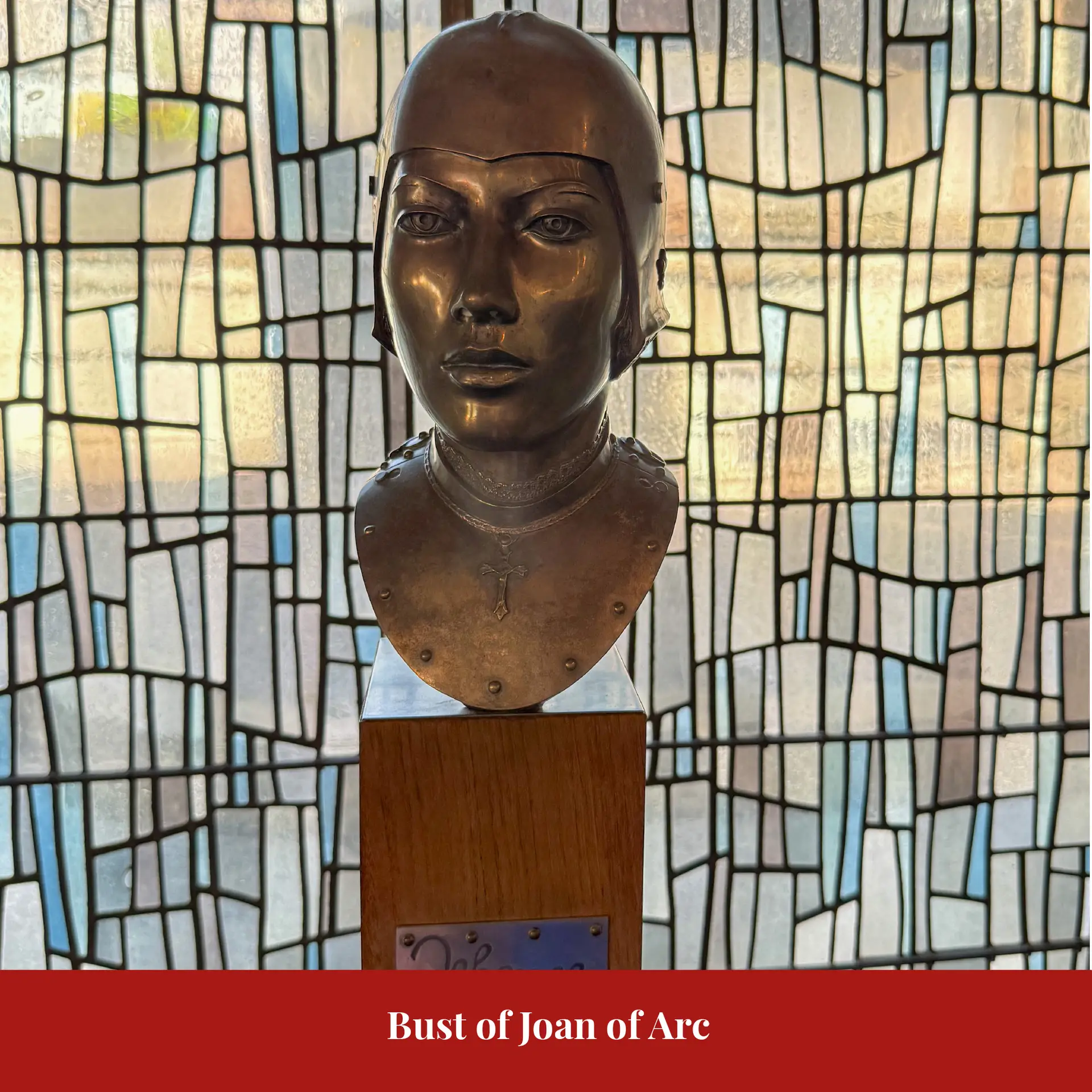 Bronze bust of Joan of Arc wearing a coif and armoured gorget with a small cross pendant, displayed on a wooden plinth against a stained-glass backdrop.
