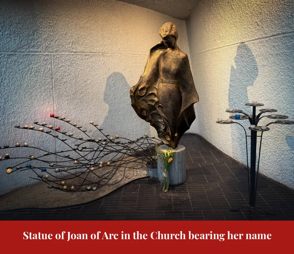 Abstract bronze statue of Joan of Arc in flowing robes, standing among rows of votive candles with flowers left at its base.