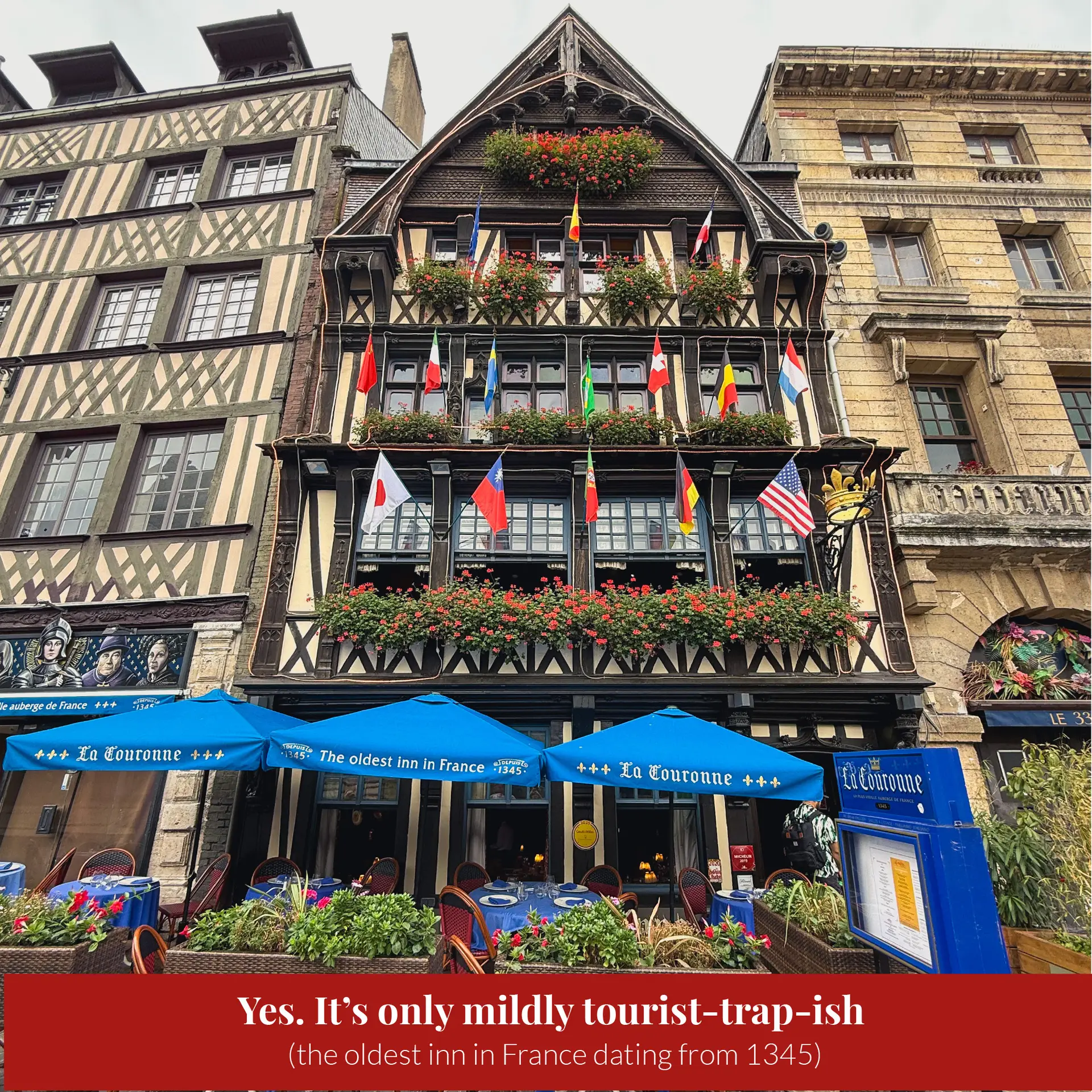 Half-timbered facade of La Couronne restaurant in Rouen, dating from 1345, decorated with international flags and flower boxes, with blue umbrellas over outdoor tables.