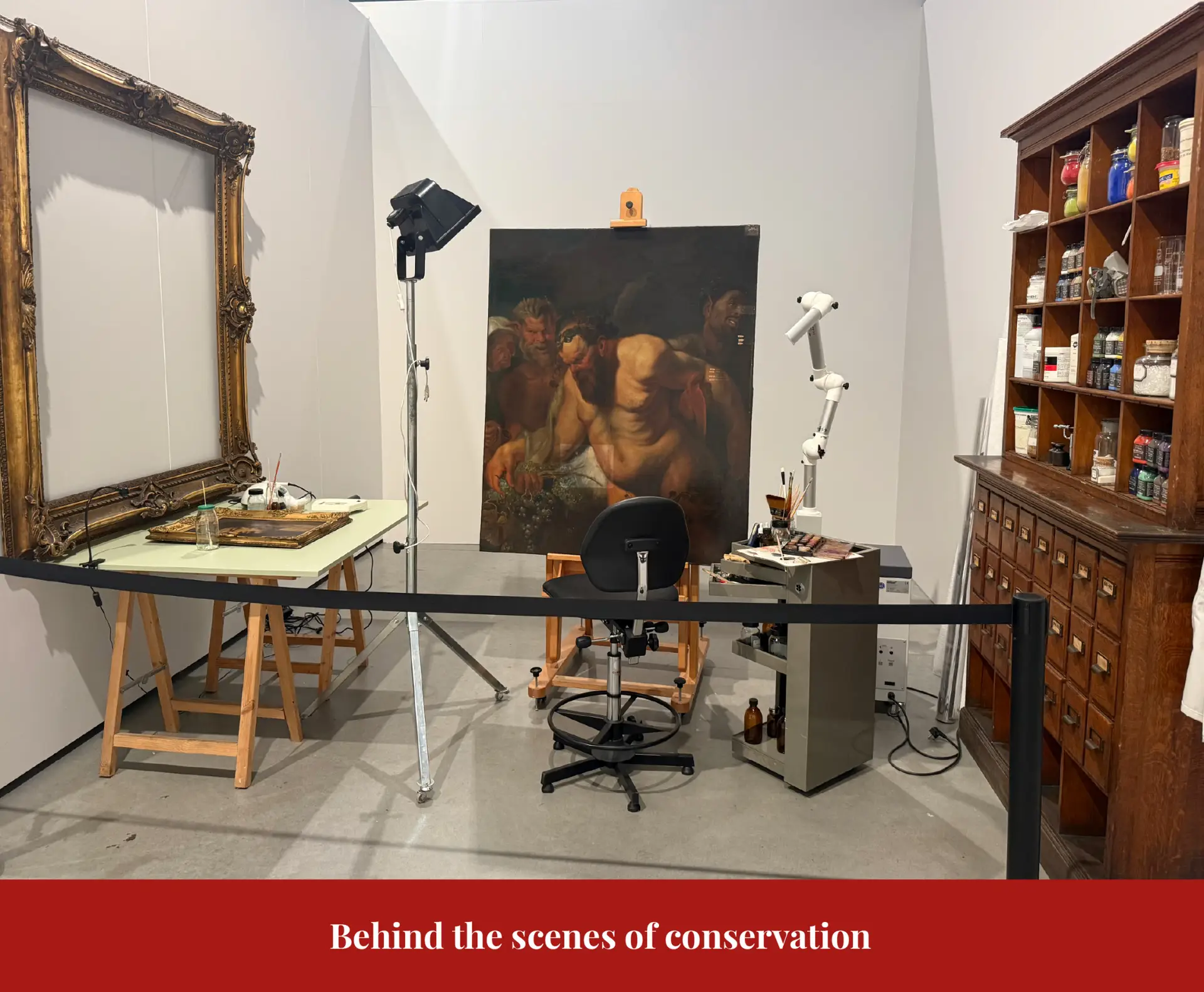 Conservation studio at La Boverie: a large painting on an easel under studio lights, beside an empty gilt frame and a cabinet of materials.